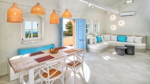 A spacious living area with a large wooden table, white chairs, a sofa with blue cushions, and a view of the outdoors through an open door. - Sienna Eco Resort (Santorini)