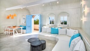 A spacious living area with a white sofa, a dining table with chairs, and a large window with a view. - Sienna Eco Resort (Santorini)