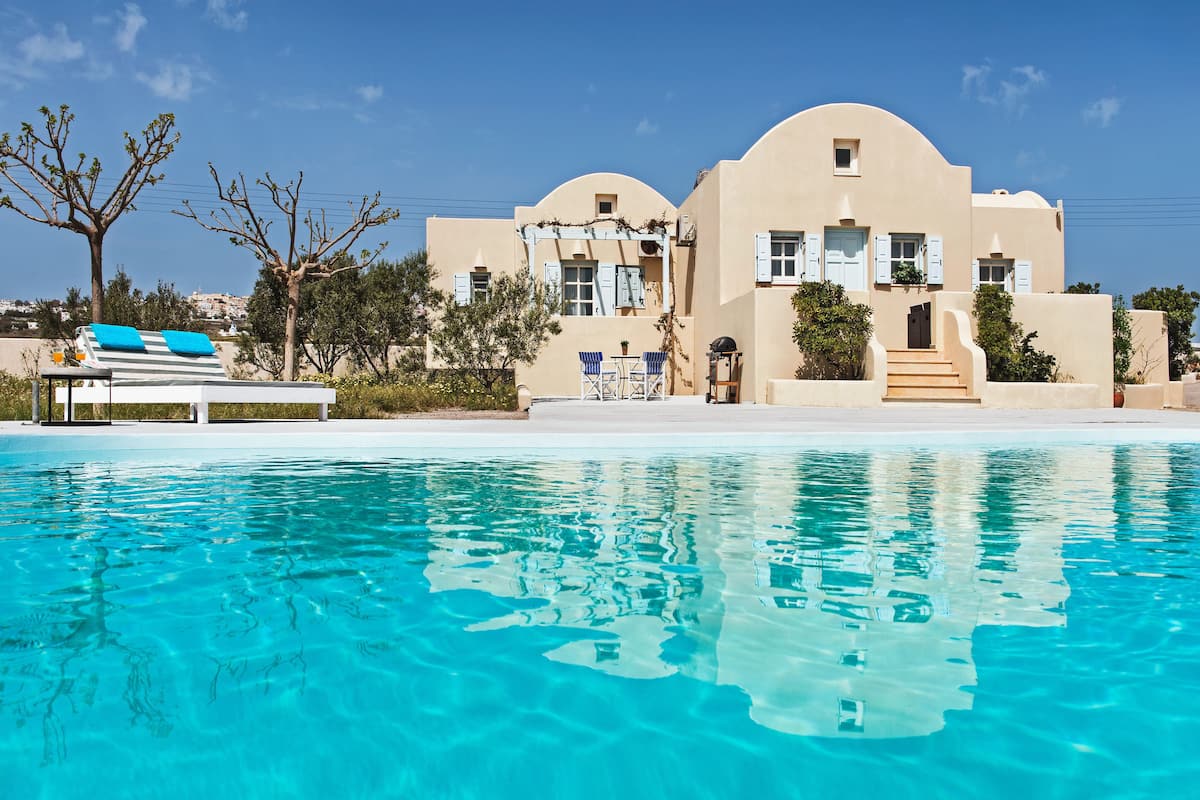 A poolside view of a Mediterranean-style building with a clear blue pool, outdoor seating, and a staircase leading to the entrance.