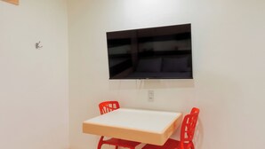 Ein kleiner Raum mit einem weißen Tisch und zwei roten Stühlen, einem flachbildfernseher an der Wand und einer Steckdose.