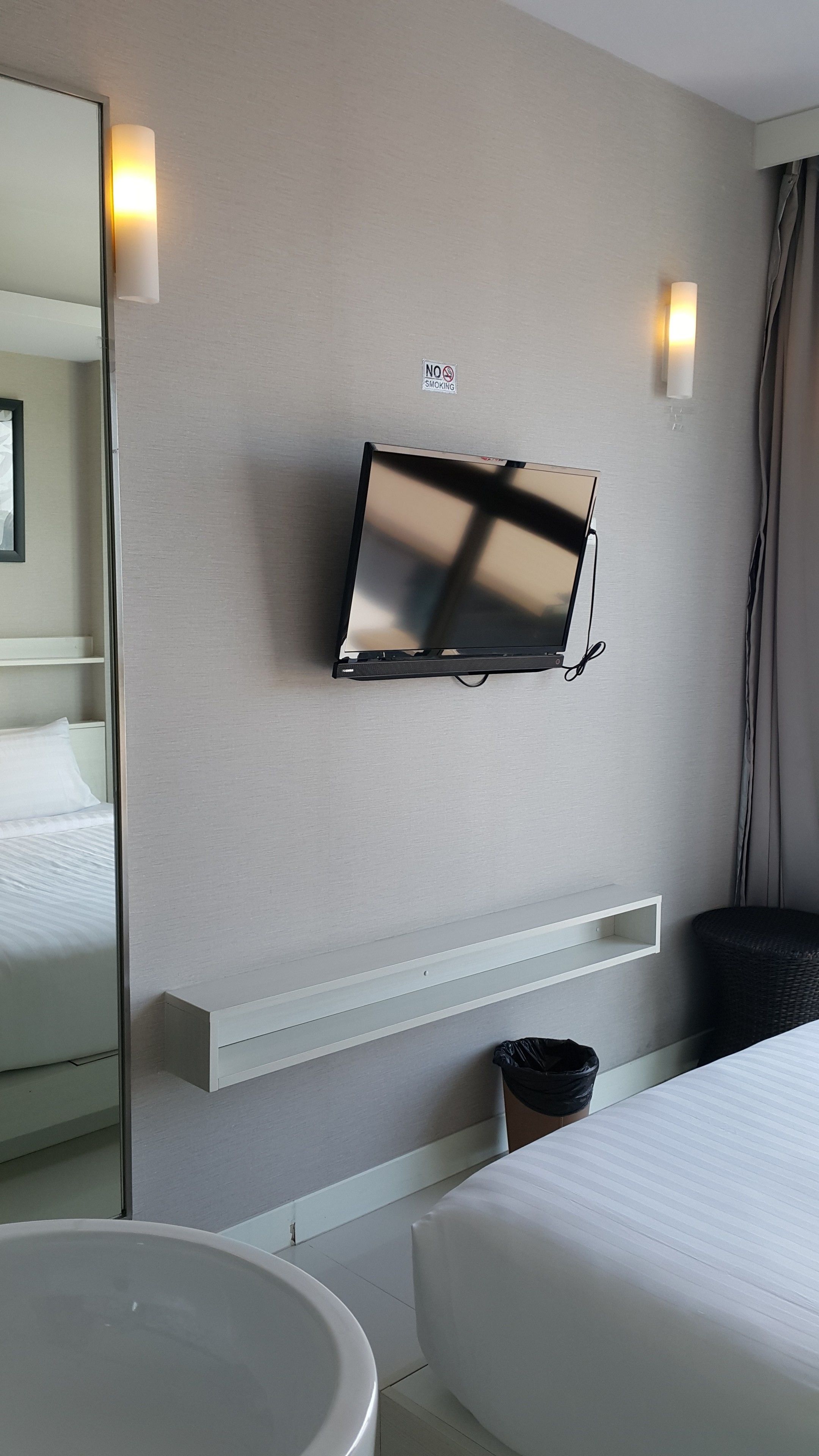 Una habitación de hotel moderna con una televisión montada, una mesita de noche, un cubo de basura pequeño y un espejo grande.