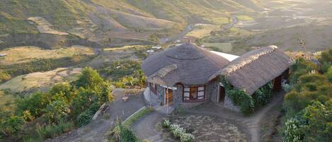 A thatched-roof house with a stone foundation, surrounded by terraced fields and a valley.