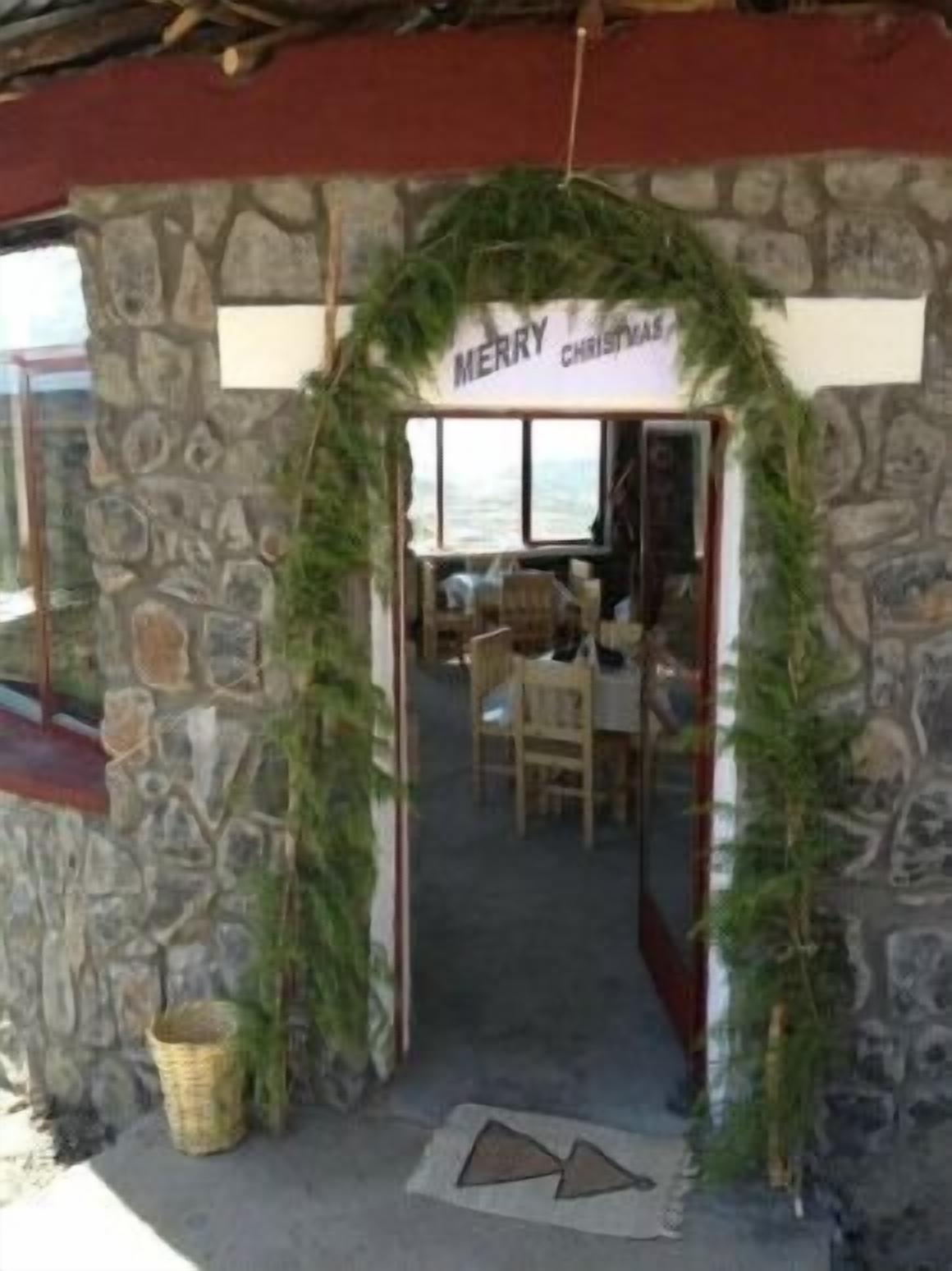 A stone building entrance with a 'Merry Christmas!' sign. Inside, there is a dining area with tables and chairs.
