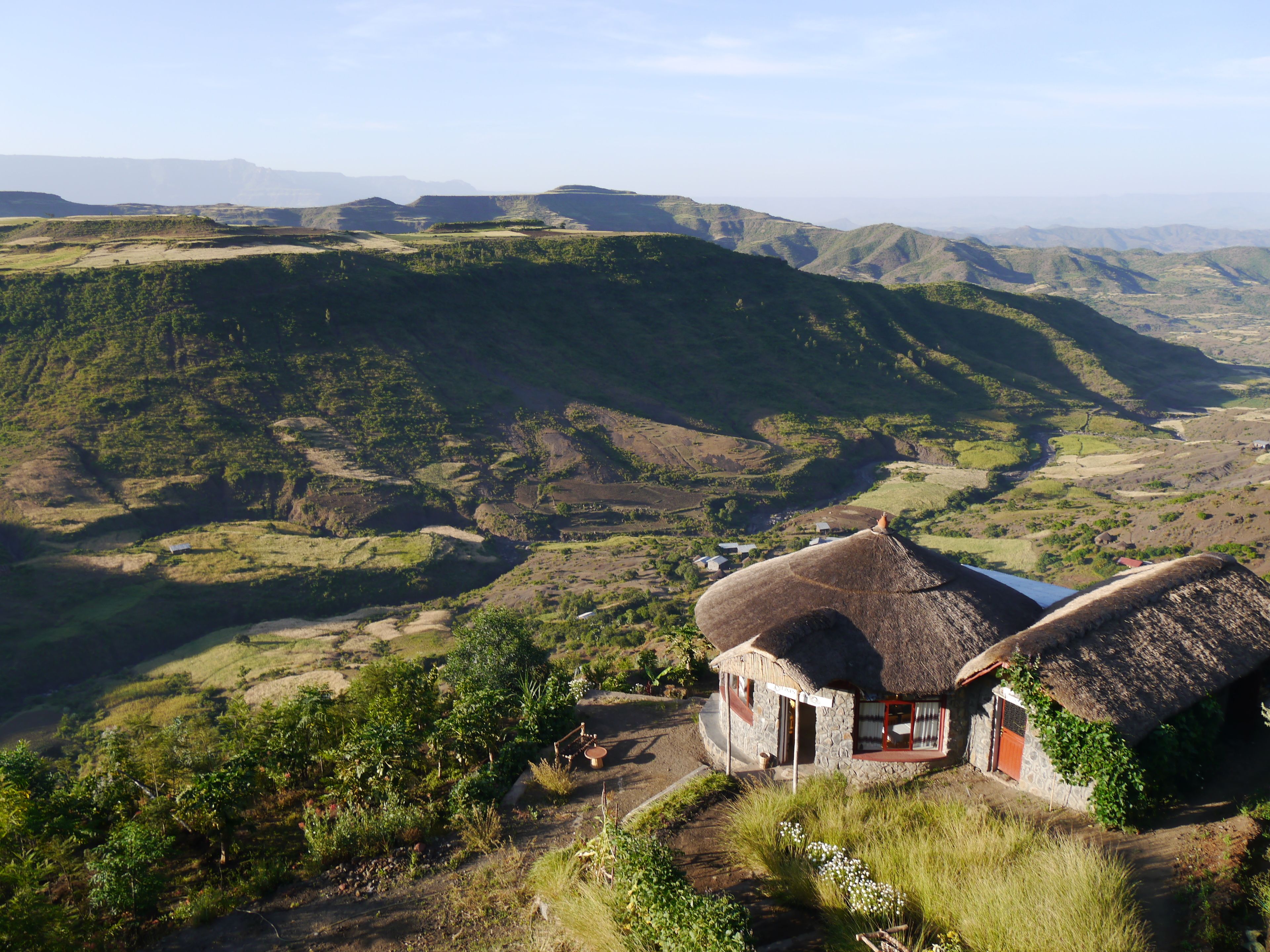 A thatched-roof house with a stone foundation, surrounded by lush green hills and a valley.