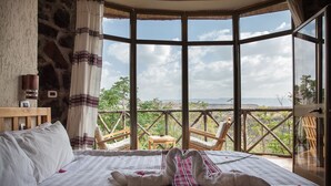 A bedroom with a large window, a bed with white linens, and a view of greenery and mountains.