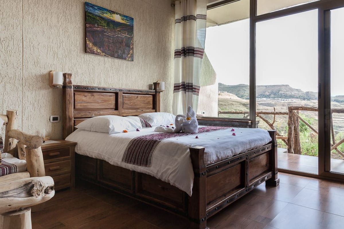 Une chambre à coucher avec un lit en bois, une table de chevet, une chaise en bois et une vue sur l’extérieur grâce à de grandes fenêtres.