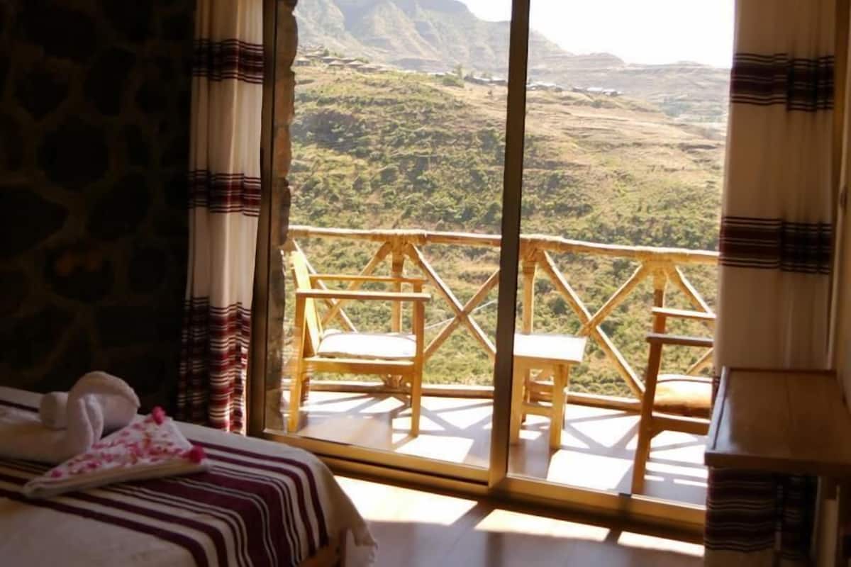 Une chambre dotée d’un lit, d’une grande fenêtre donnant sur un paysage montagneux et d’un balcon agrémenté de meubles en bois.