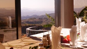 A dining table set with white napkins, salt and pepper shakers, and a view of a mountainous landscape through large windows.