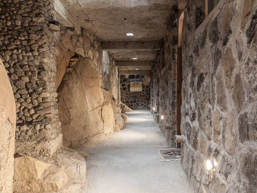 Un couloir étroit aux murs de pierre et au sol en béton, qui mène à une pièce dont un des murs est également en pierre et qui possède une fenêtre.