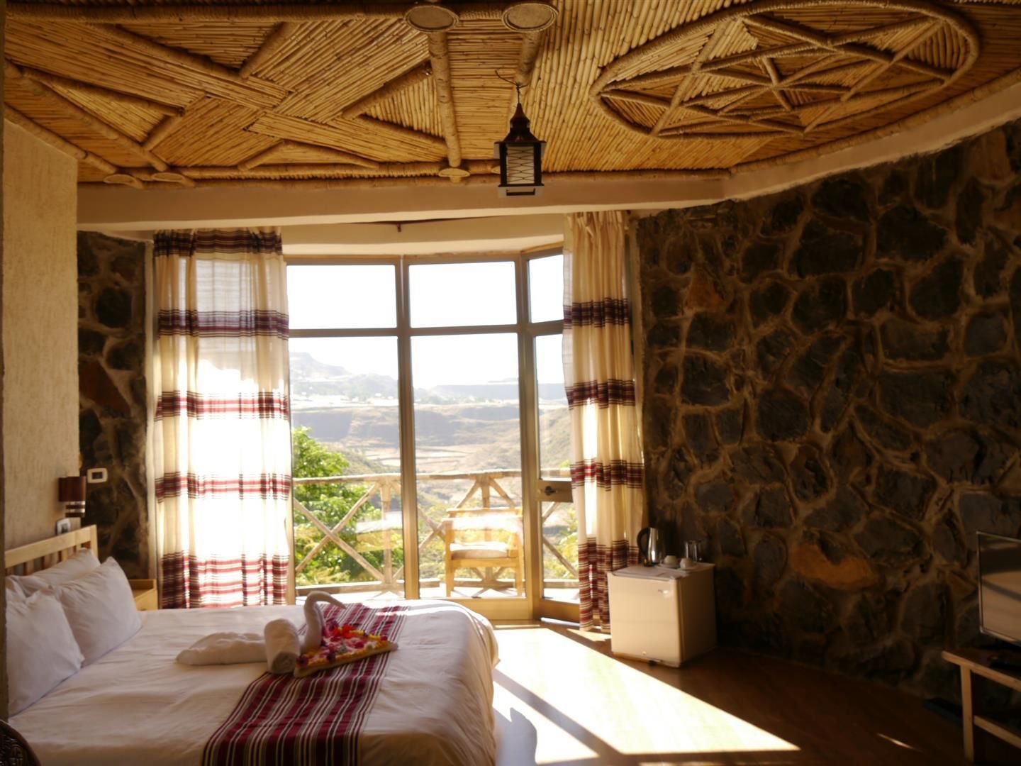 Una habitación con cama, ventana con cortinas, pared de piedra y una mesa con hervidor.