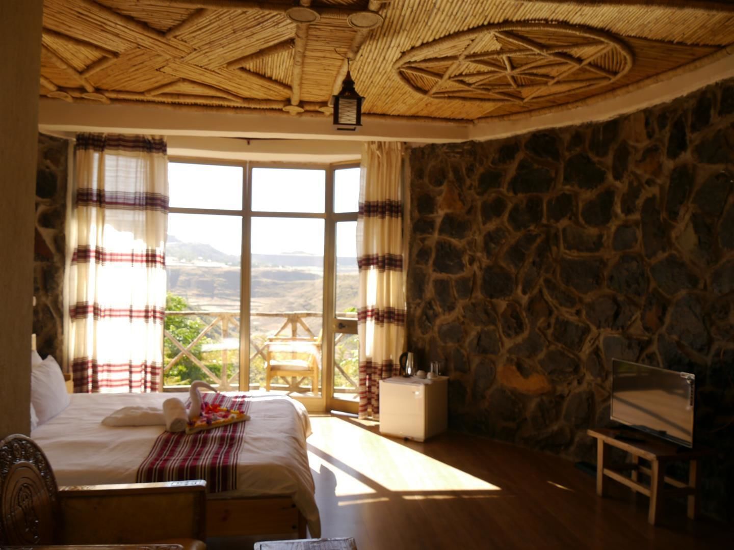 Quarto com parede de pedra, uma cama com manta xadrez, mesa de cabeceira de madeira e vista para o exterior.