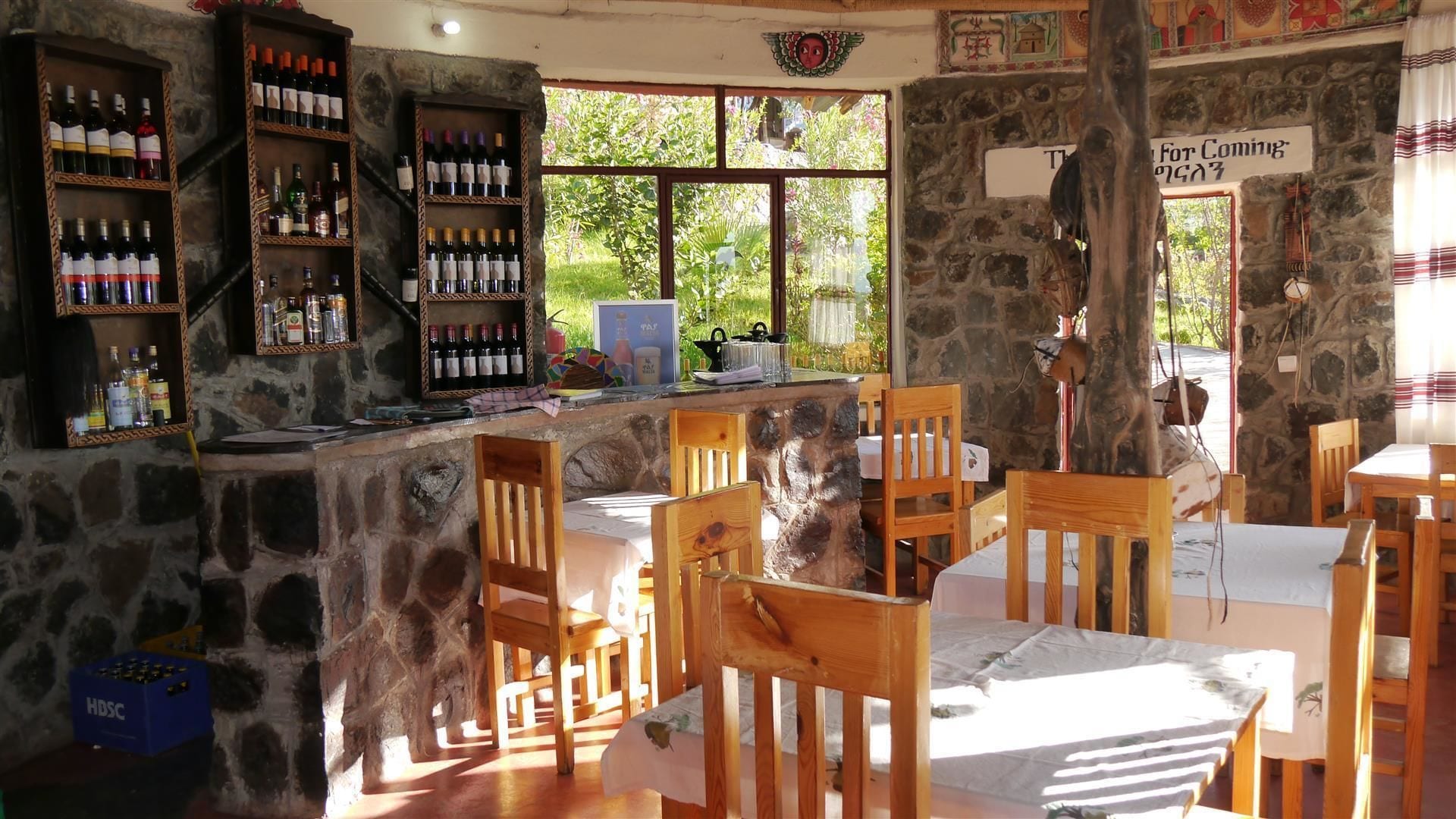 Un área de bar rústica con estantes de madera llenos de botellas, paredes de piedra y vistas al exterior con vegetación.