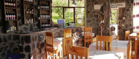 Un área de bar rústica con estantes de madera llenos de botellas, paredes de piedra y vistas al exterior con vegetación.