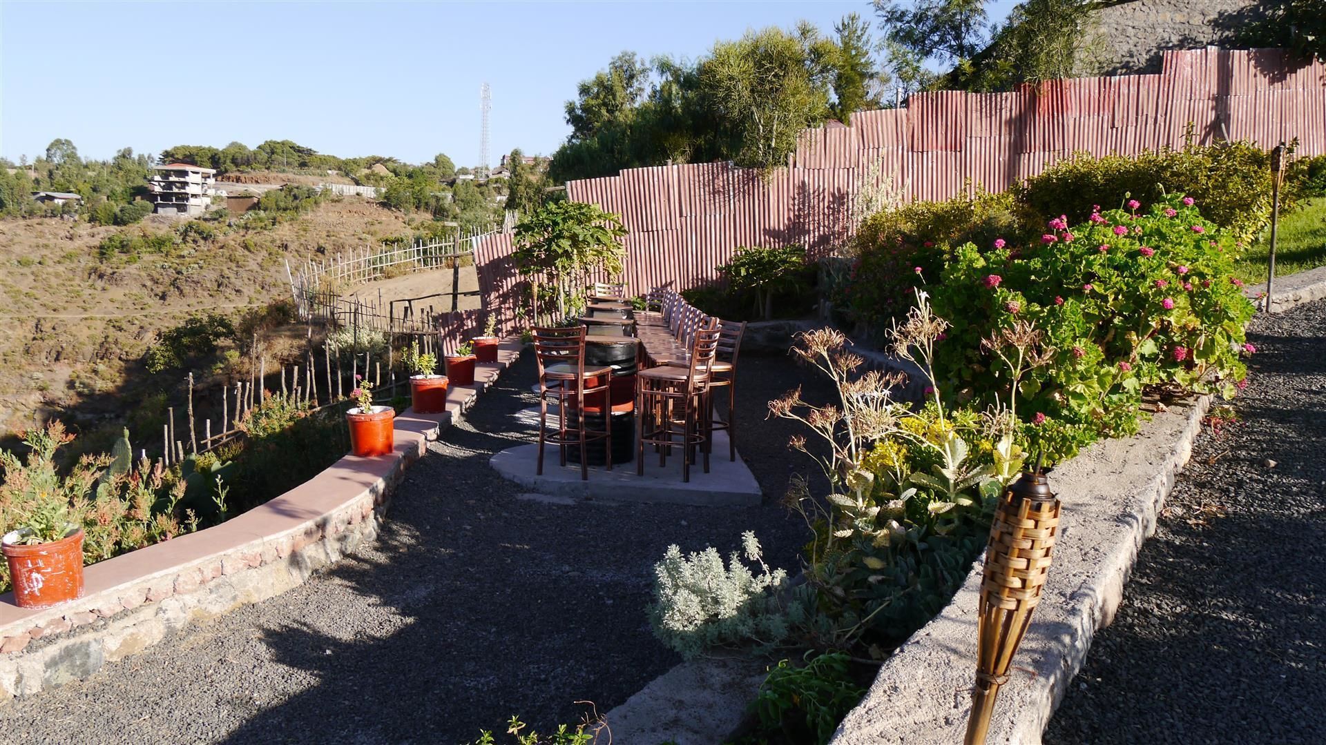 Un patio exterior con una mesa larga y sillas, rodeado de plantas en macetas y una valla de madera.