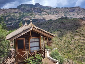 A wooden cabin with a thatched roof, a balcony, and a view of a mountainous landscape.