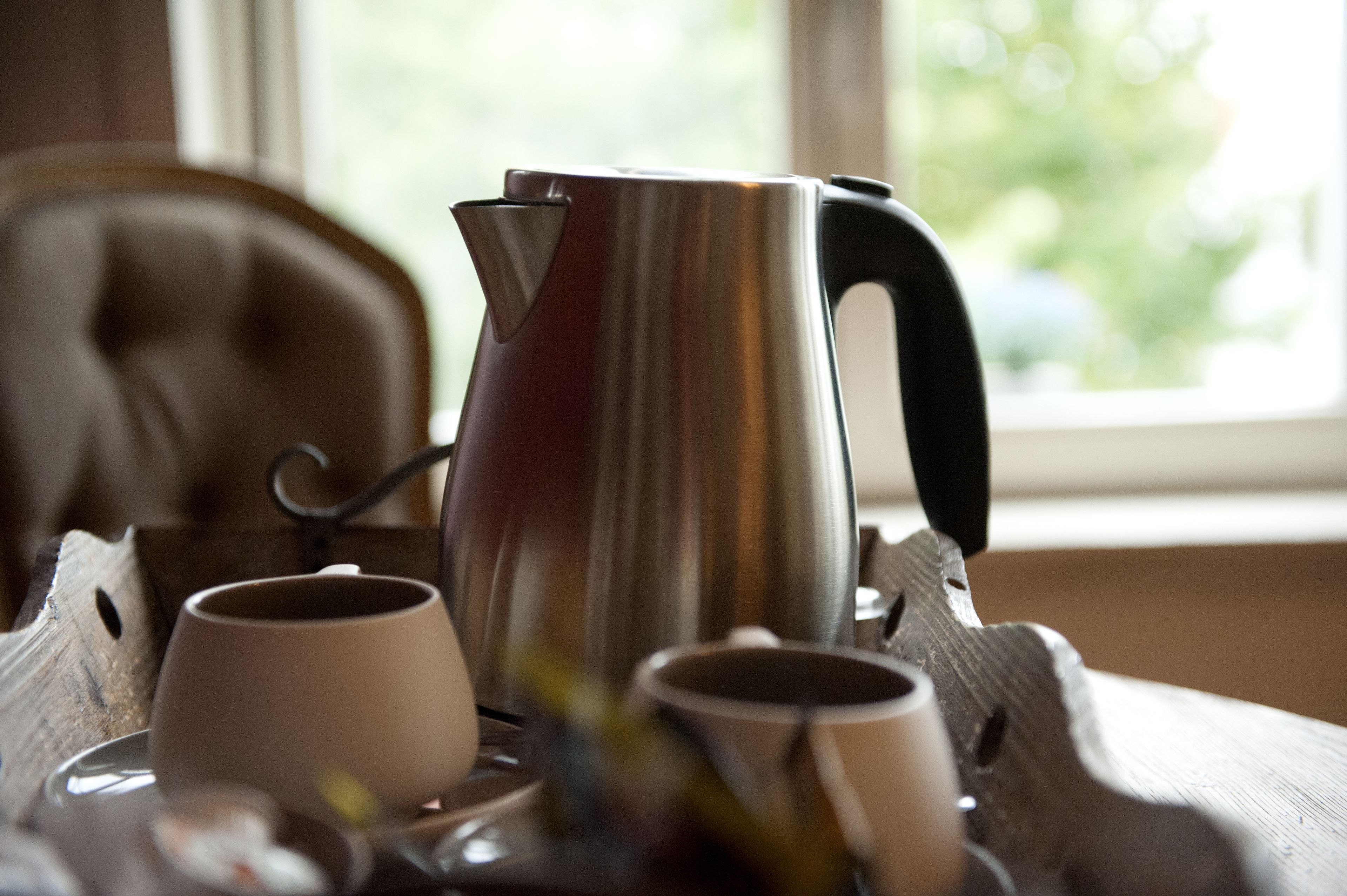 Una cafetera de acero inoxidable sobre una bandeja con dos tazas, colocada sobre una mesa de madera cerca de una ventana.