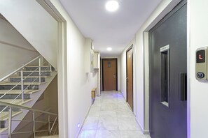 Un corridoio d'hotel con una scala a sinistra, una porta con un cartello a destra e un ascensore con il pannello di controllo.