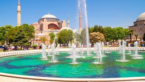Un'ampia fontana con numerosi getti d'acqua al centro, circondata da una vasca di acqua limpida. Sullo sfondo, si vedono diversi edifici con cupole e minareti, e alberi che costeggiano l'area.