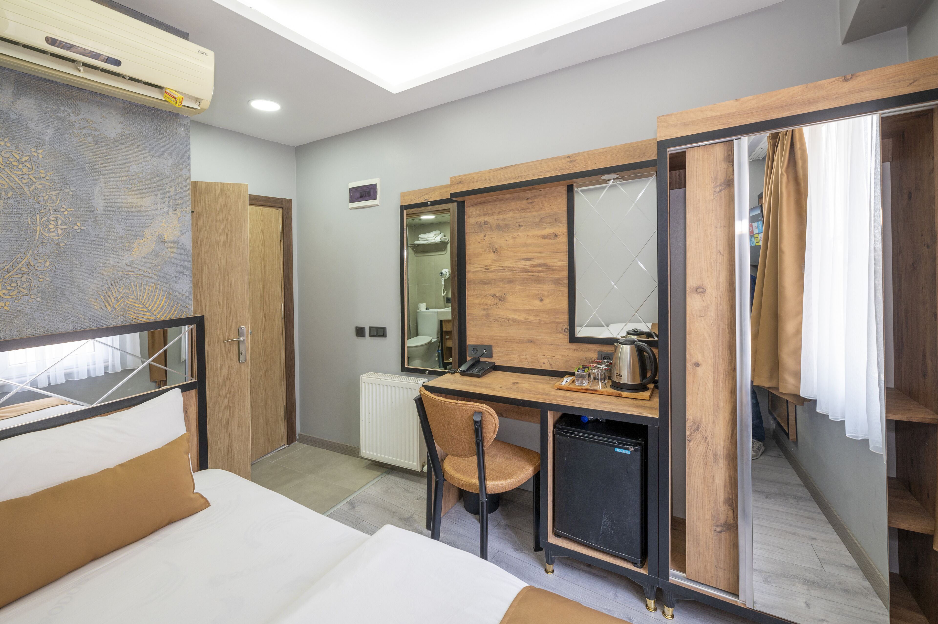 A hotel room with a bed, a desk with a chair, a small kitchenette, a mirror, and a wardrobe.