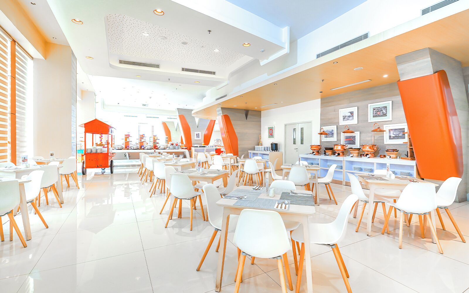 A spacious dining area with white tables and chairs, a food cart, and a kitchen area with pots and pans.