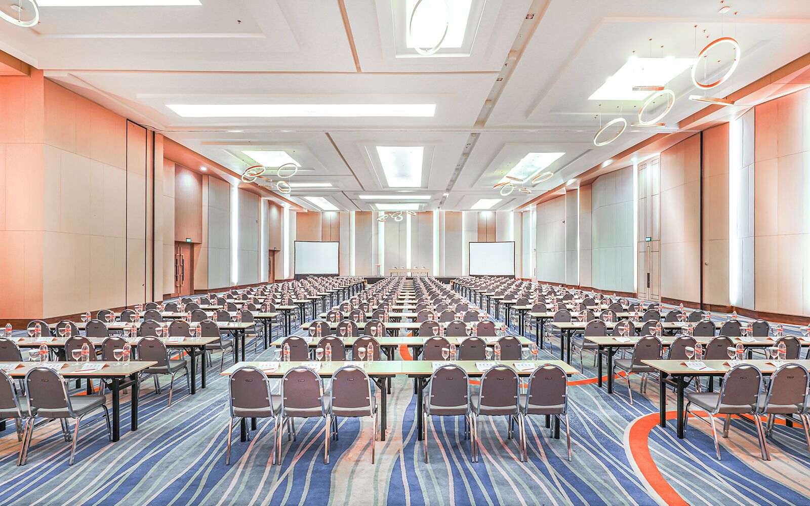 A conference room with rows of tables and chairs, equipped with water bottles and papers, and large screens at the front.