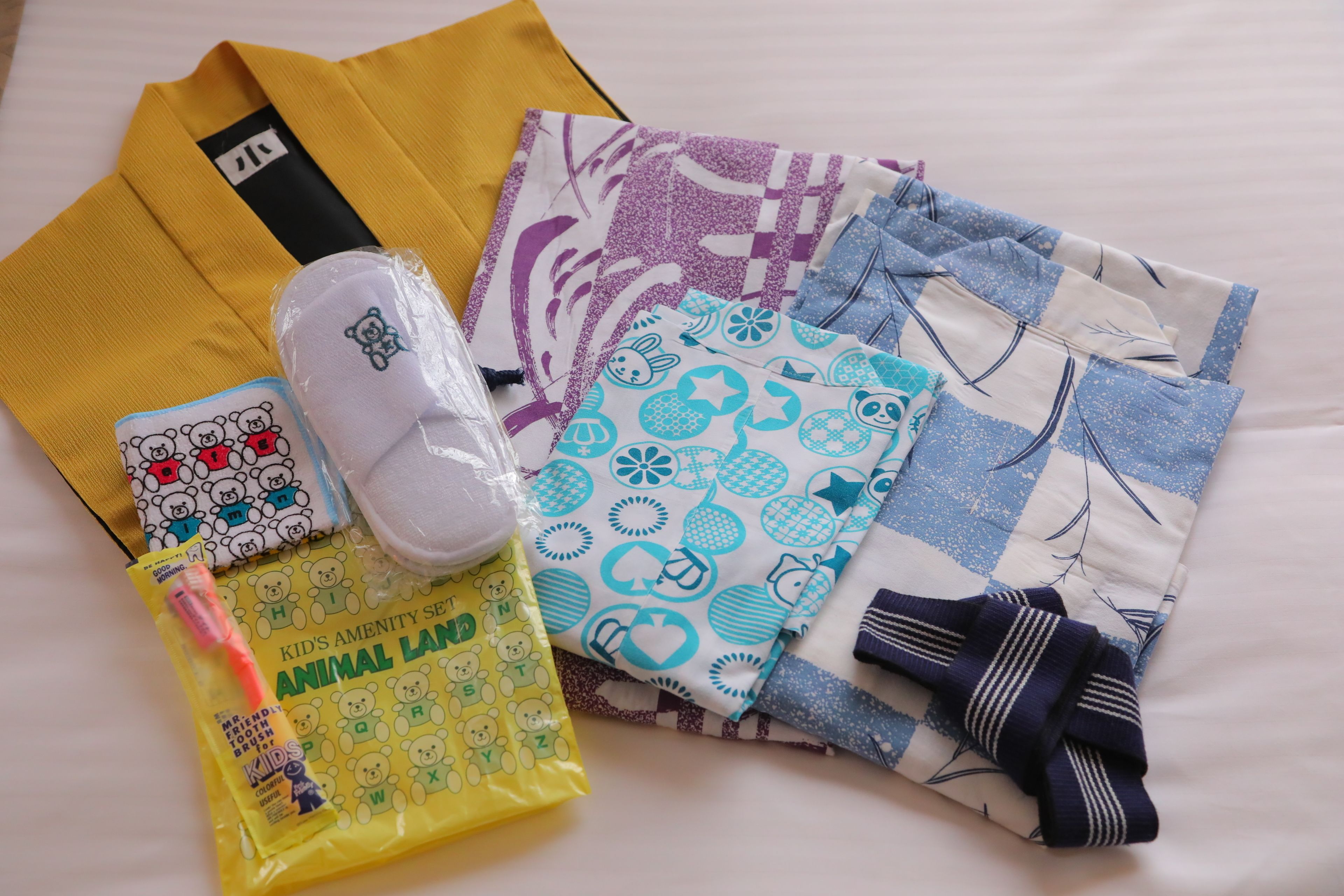 Una cama con un edredón amarillo, una almohada blanca con un diseño de oso, una almohada con estampado azul y blanco, y un bolso amarillo con un diseño de oso.