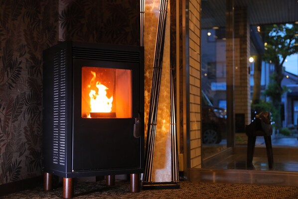 A wood-burning stove with visible flames, positioned against a patterned wallpaper and a glass door reflecting an outdoor scene.