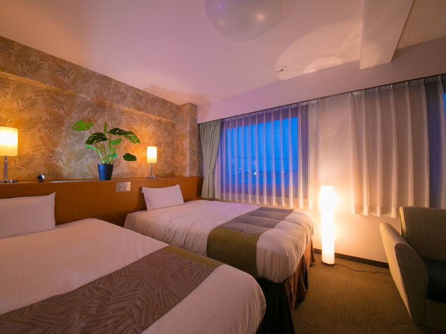 A hotel room with two beds, a potted plant, a lamp, and a view of the city at dusk.