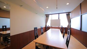 Uma sala de conferências com uma mesa comprida, cadeiras, um quadro branco e grandes janelas com cortinas.