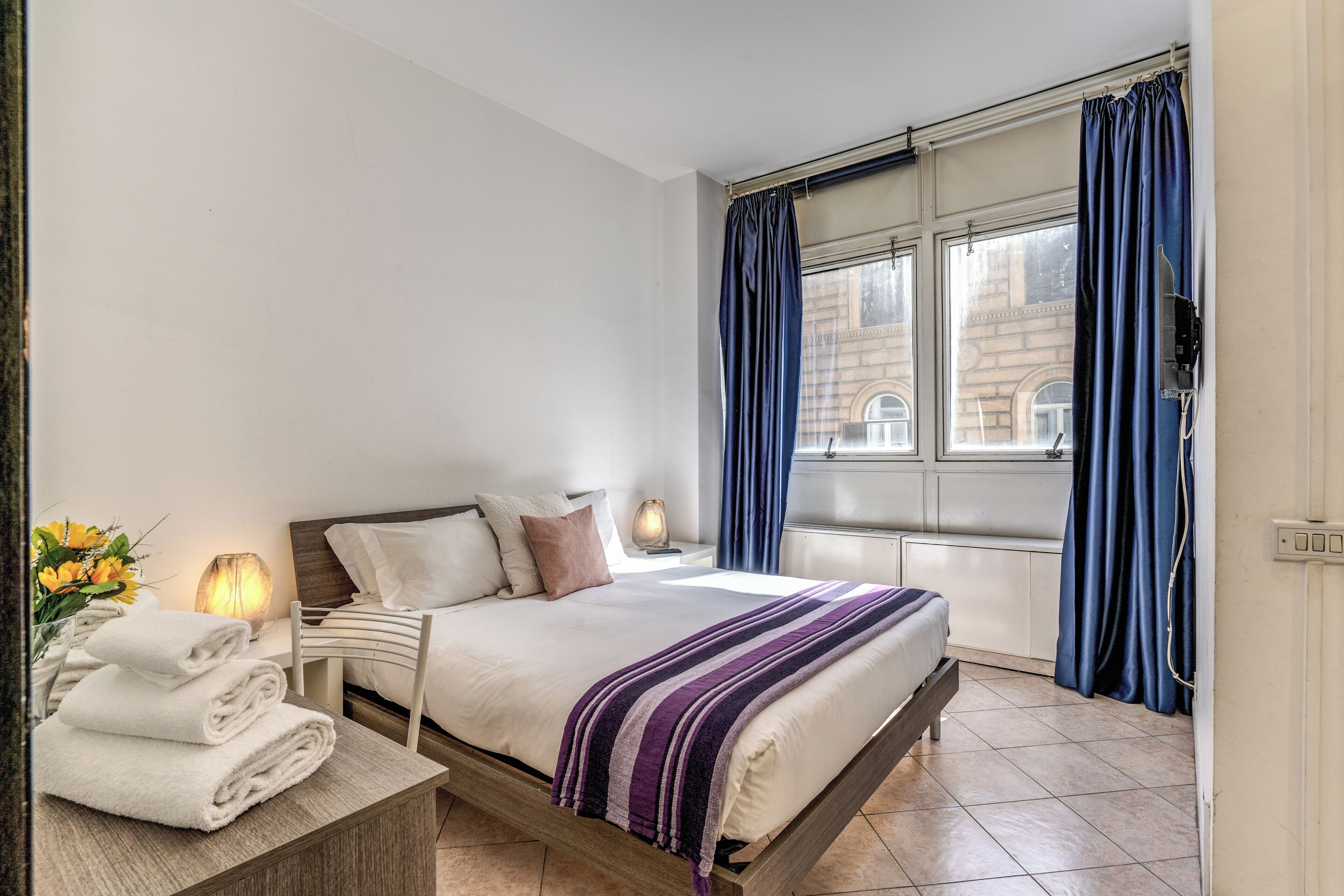 A hotel room with a bed, a bedside table with towels, a lamp, and a window with curtains.