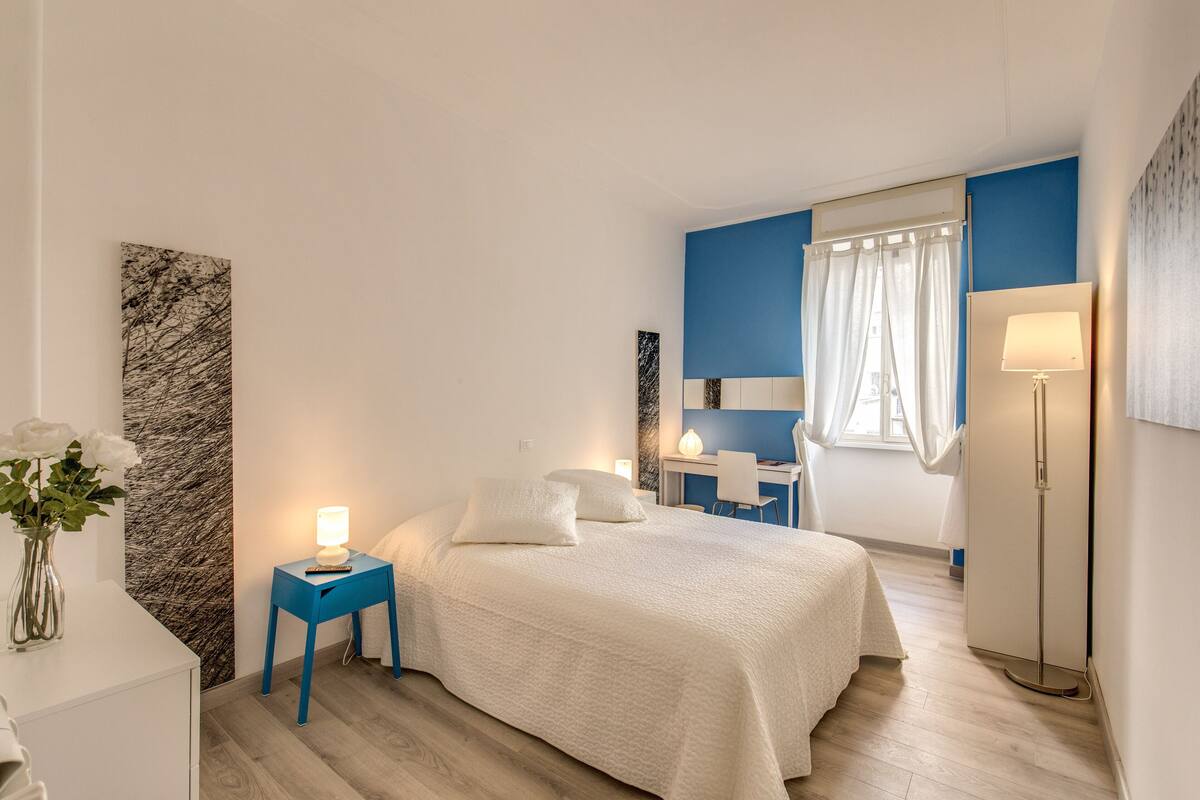 A bedroom with a bed, bedside table, lamp, desk, chair, and a large painting on the wall.