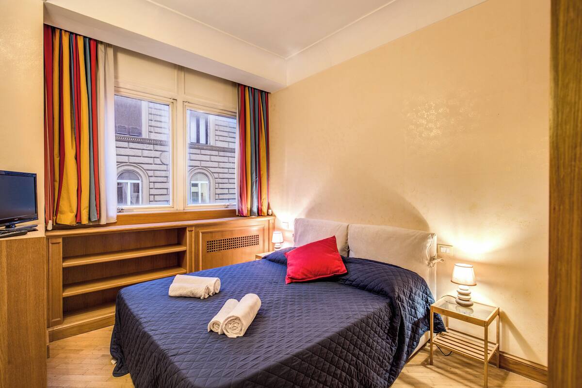 A hotel room with a bed, a TV, a desk, and a window with curtains.
