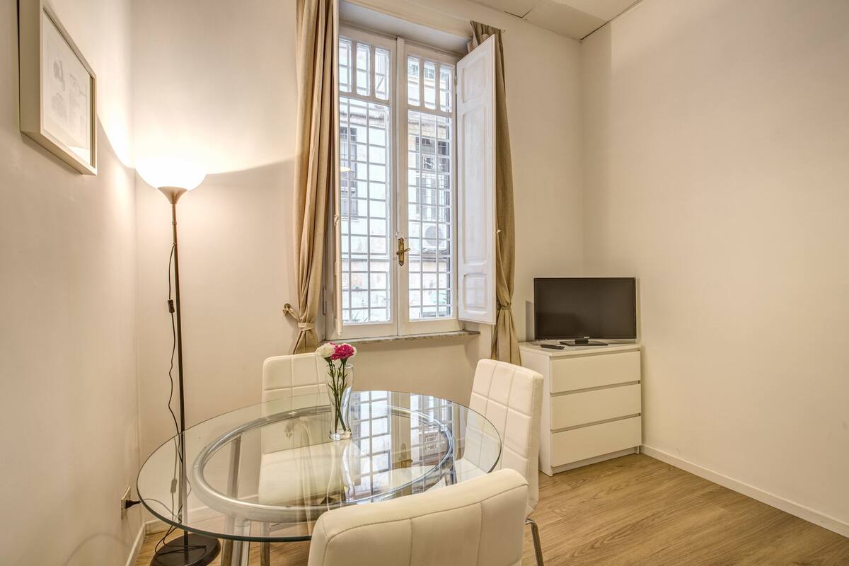 A small, well-lit room with a glass table, two chairs, a TV on a stand, and a window with curtains.