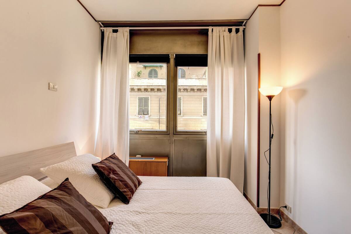 A hotel room with a bed, a nightstand, a lamp, and a view of the street through the window.