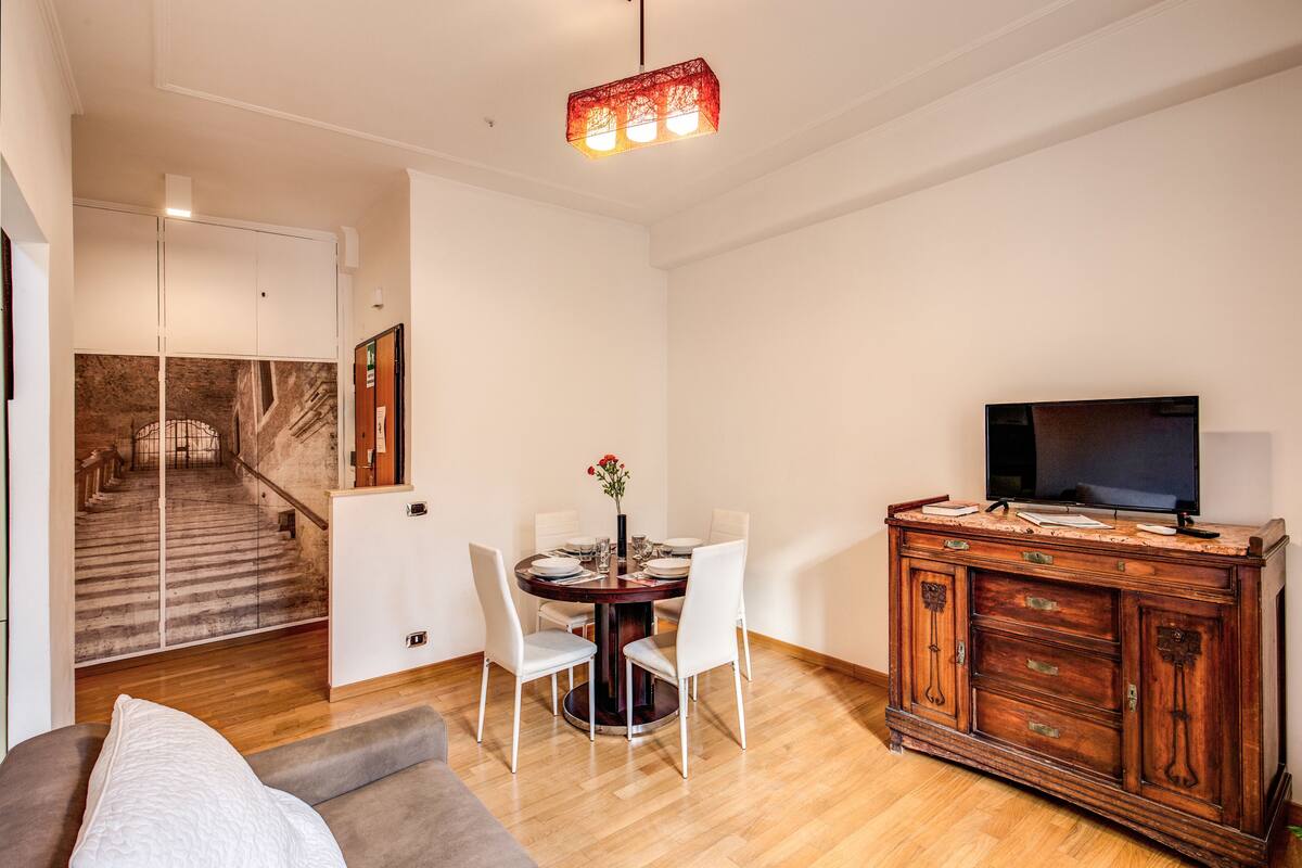 A dining area with a round table, white chairs, a wooden cabinet with a television, and a staircase in the background.