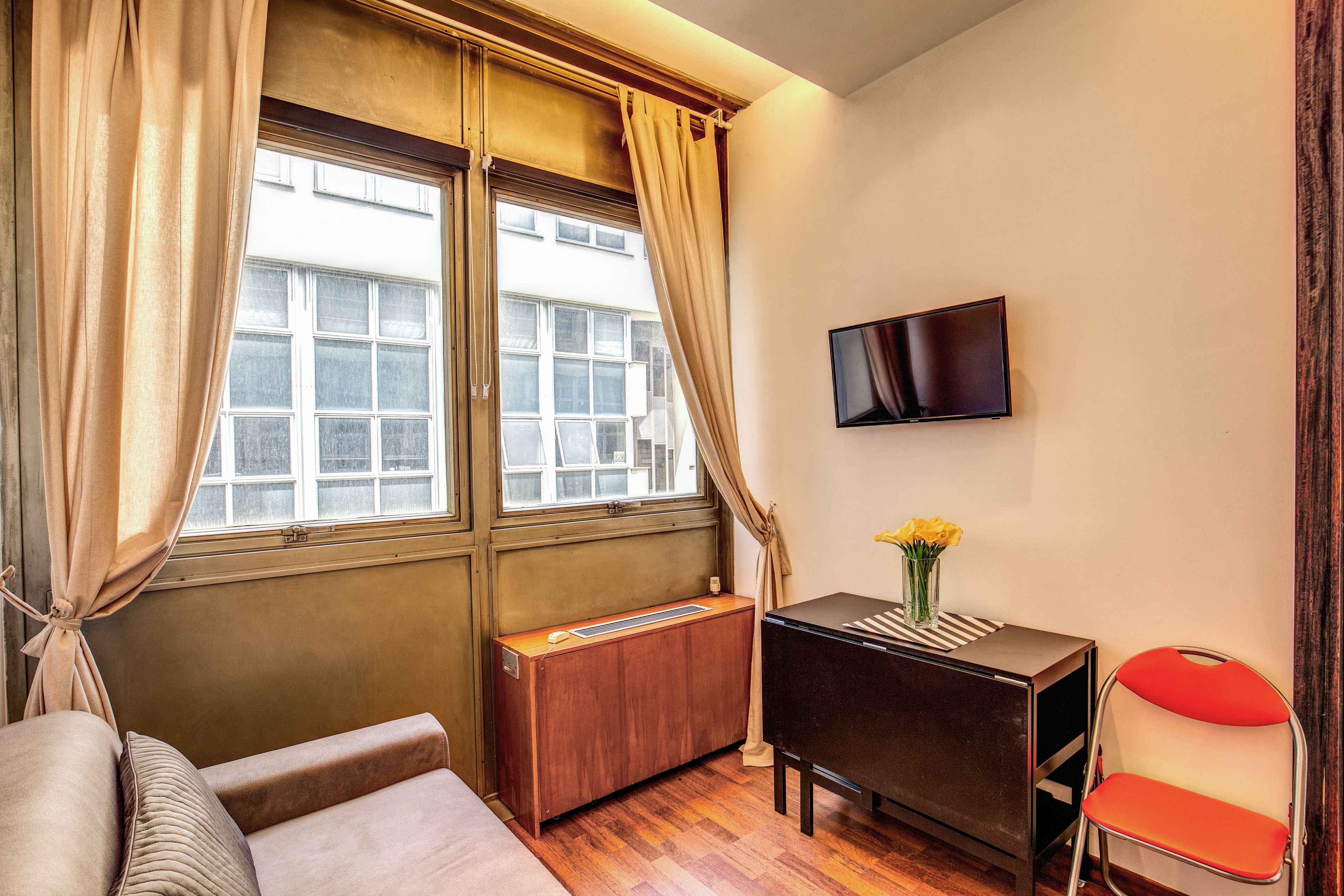 Una habitación con sofá, una mesita con un jarrón de flores, televisor montado en la pared y ventana con cortinas.