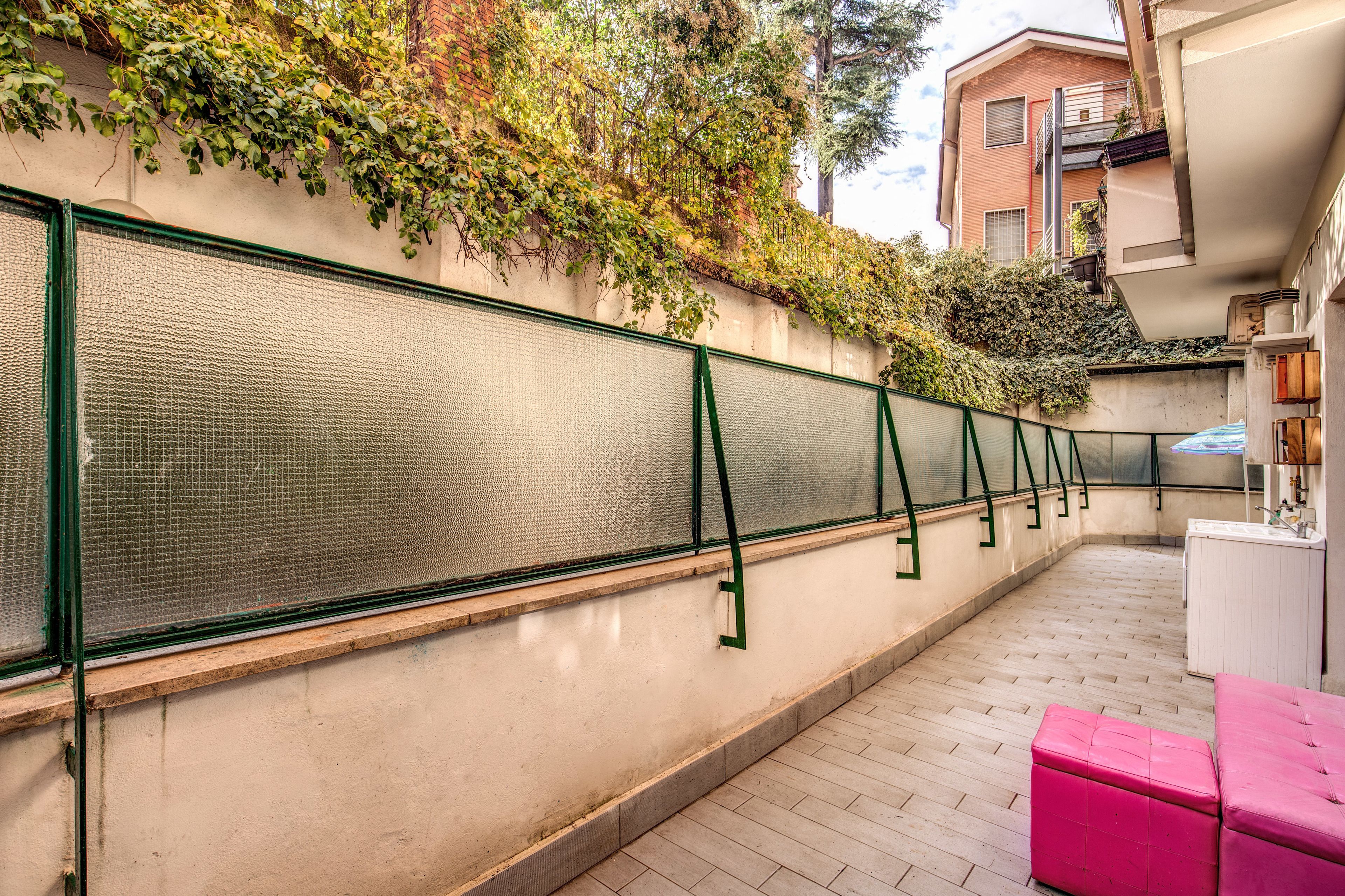 A narrow outdoor corridor with a textured wall, a green metal railing, and a pink bench.