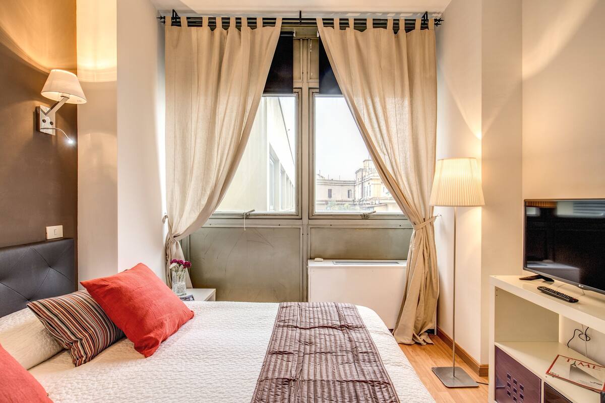 A hotel room with a bed, a TV, a lamp, and a window with curtains.