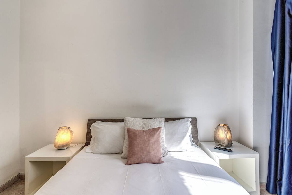 A neatly made bed with white and pink pillows, two bedside tables with lamps, and a blue curtain on the right.