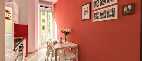 Un comedor con pared roja, mesa blanca y sillas. Hay cuadros en la pared y un jarrón con flores sobre la mesa.