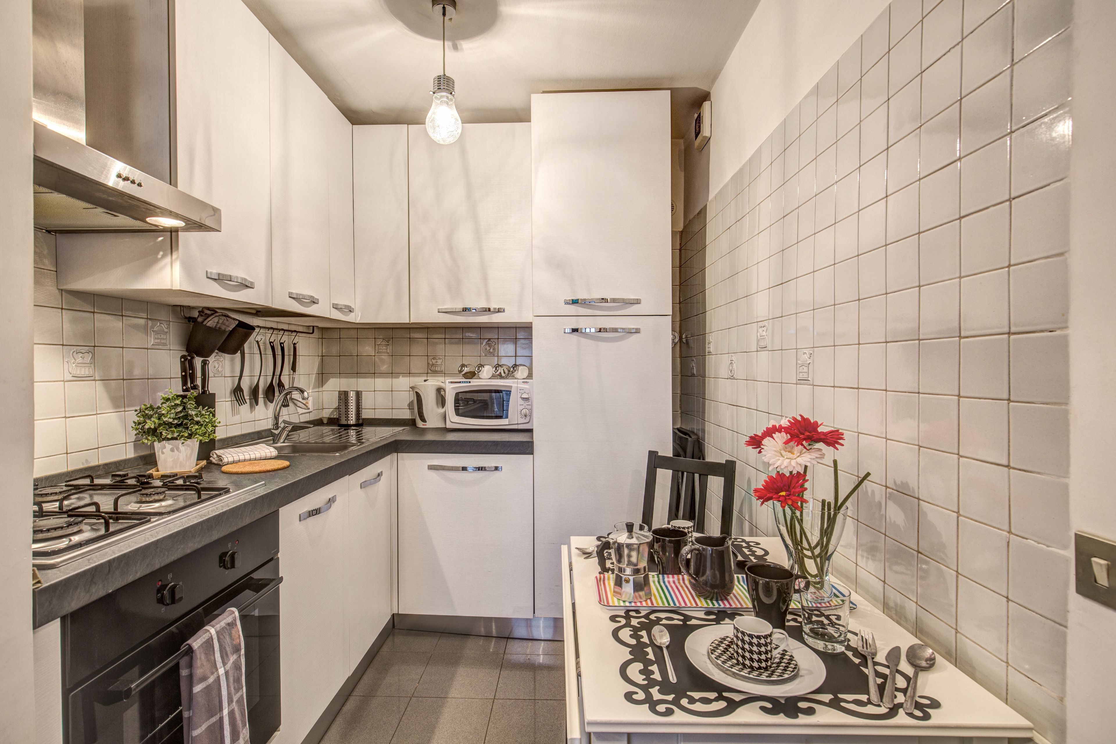 Uma cozinha compacta com armários brancos, fogão a gás, micro-ondas e um espaço de jantar com mesa posta para dois.