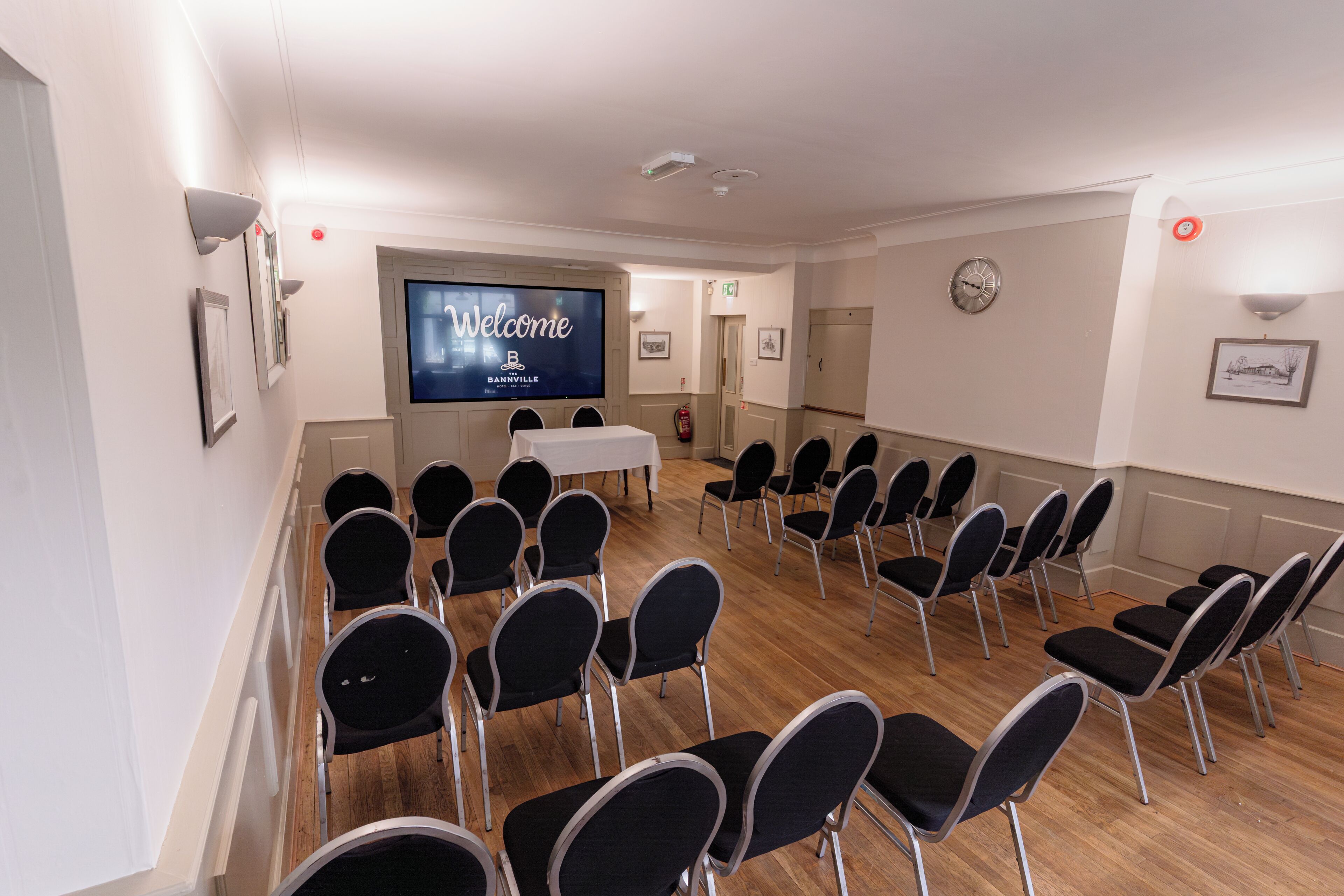 Un salón de conferencias con filas de sillas, una mesa blanca, una pantalla que muestra 'Welcome B&B Barnville' y cuadros enmarcados en las paredes.