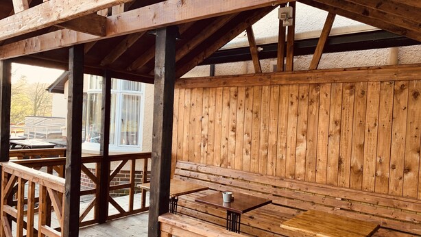 Een terras van hout buiten, met banken en tafels, een uitzicht op een gebouw met ramen en een betegelde vloer van baksteen.