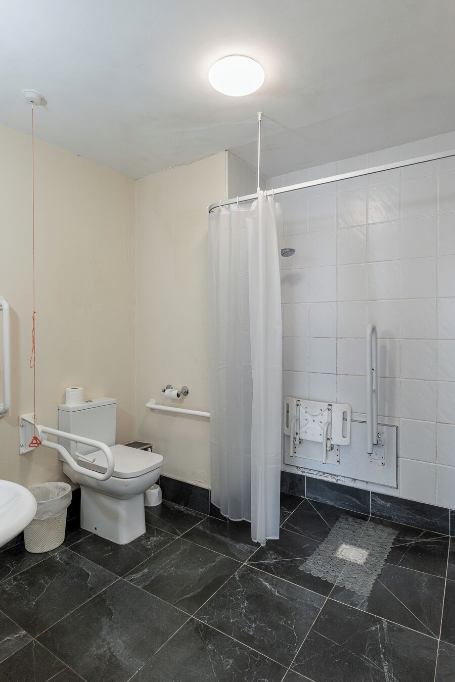Un baño con inodoro, ducha con cortina, barandilla y suelo de baldosas.