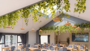 Een ruime vergaderzaal met meerdere tafels en stoelen, een groot scherm waarop 'Welkom' staat en hangplanten.