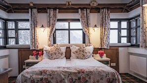 Un dormitorio con una cama grande, ropa de cama floral, dos mesitas de noche con lámparas y vistas a árboles nevados a través de las ventanas.