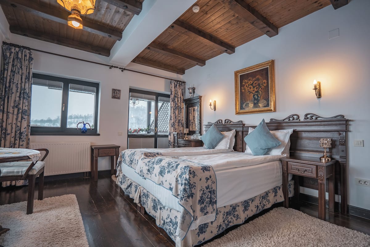Quarto com teto de madeira, uma cama grande com roupa de cama floral, duas mesas de cabeceira e uma janela com cortinas.