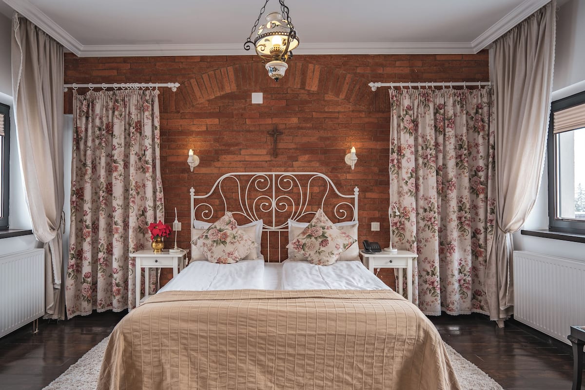 Quarto com cabeceira de madeira, cortinas florais, cama com colcha bege, mesas de cabeceira com abajures e vista pela janela.