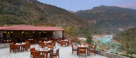 An outdoor restaurant with wooden tables and chairs, set against a backdrop of mountains and a river.