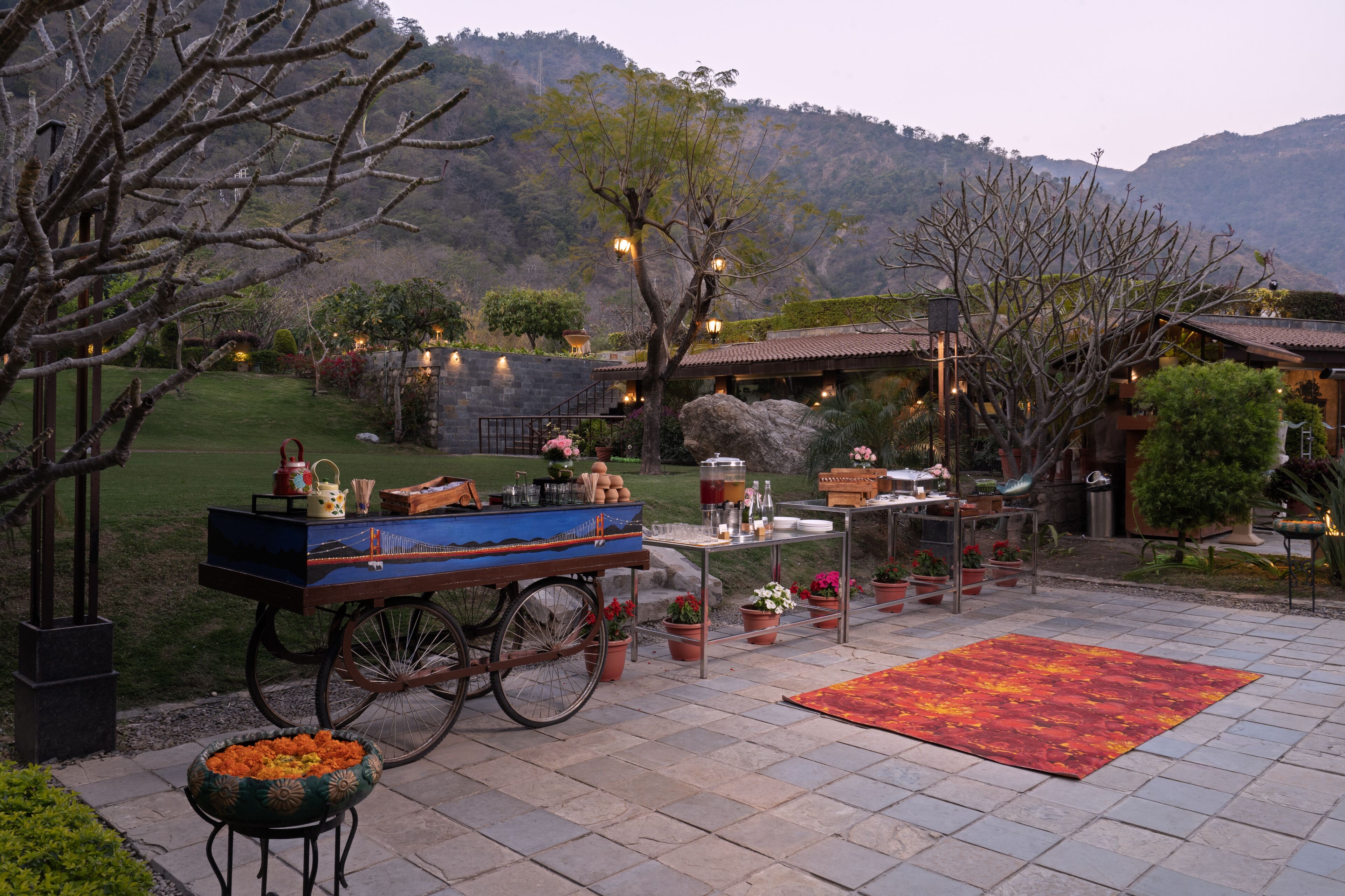 A patio with a cart, table, and chairs, surrounded by potted plants and a view of mountains.